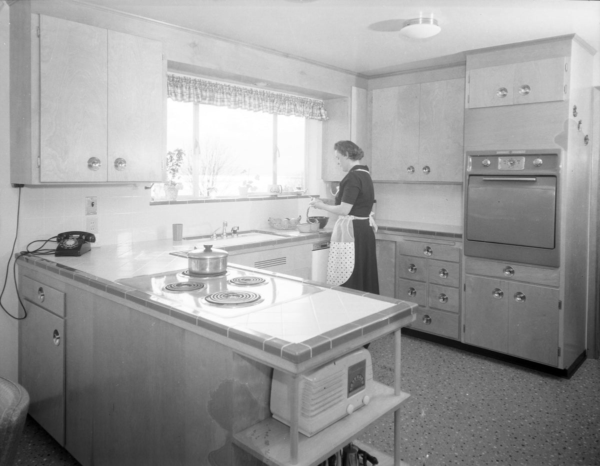 A woman in a 1956 all-electric kitchen with an island cooktop and a built-in wall oven